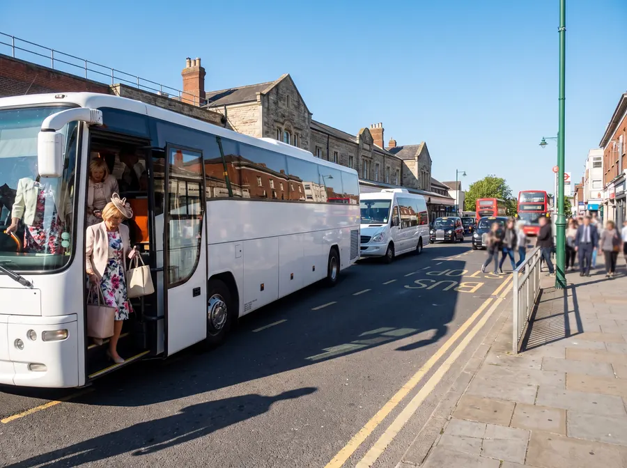 A white touring coach with a minibus parked behind it on the road outside Sutton railway station in bright summer sunlight, a group of women dressed up for a day out boarding with bags